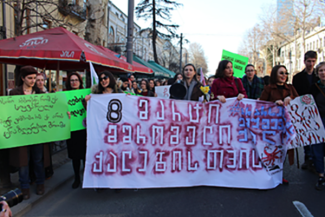 The photo shows a demonstration where a crowd of people is holding a banner in Georgian that reads as follows: “8th of March for worker women”.