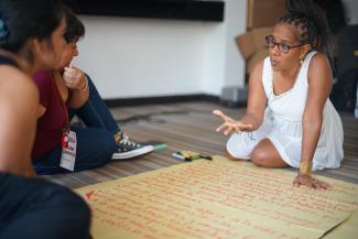 Two women sitting on the floor in conversation during a group activity. One of them, wearing a white dress and glasses, is gesturing expressively while the other listens attentively. A large sheet of paper with handwritten red text is placed between them.